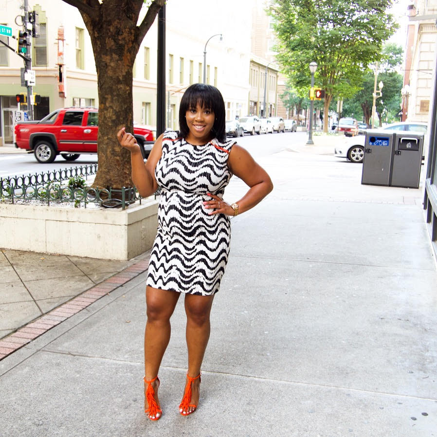 A black woman smiling, wearing a black and white dress, standing on street with hand on hip